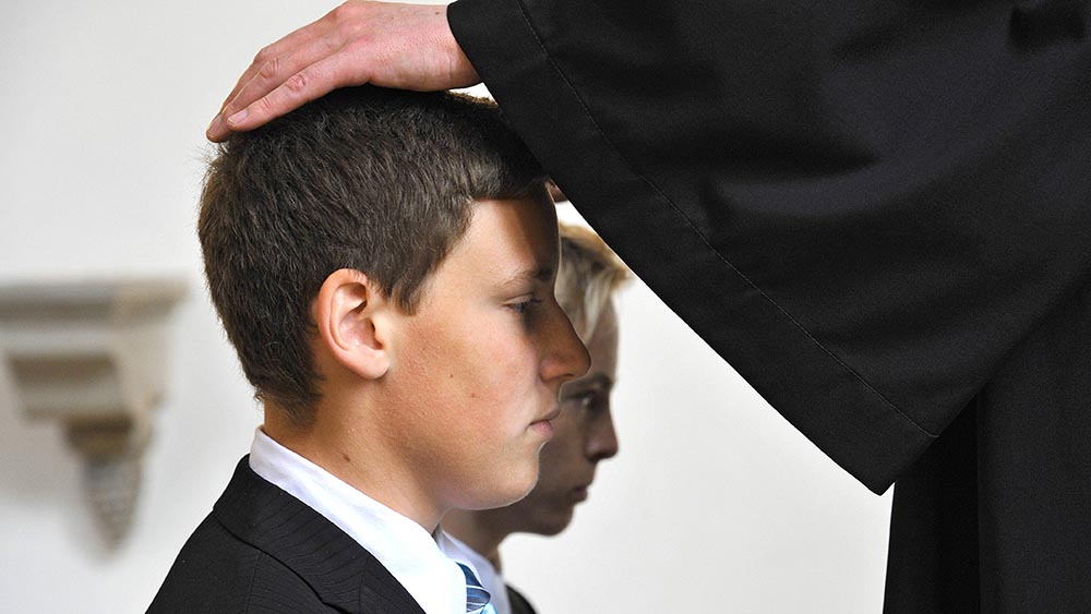 Jugendliche im Anzug knien vor Altar. Pastor hält Hand zum Segen auf den Kopf