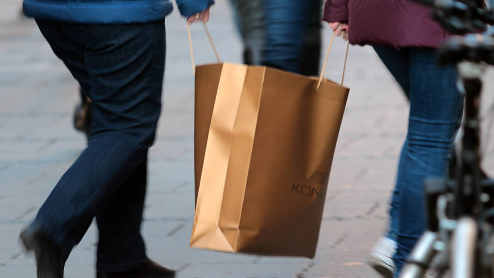 Passanten tragen gemeinsam eine volle und schwere Einkaufstasche durch eine Fußgängerzone