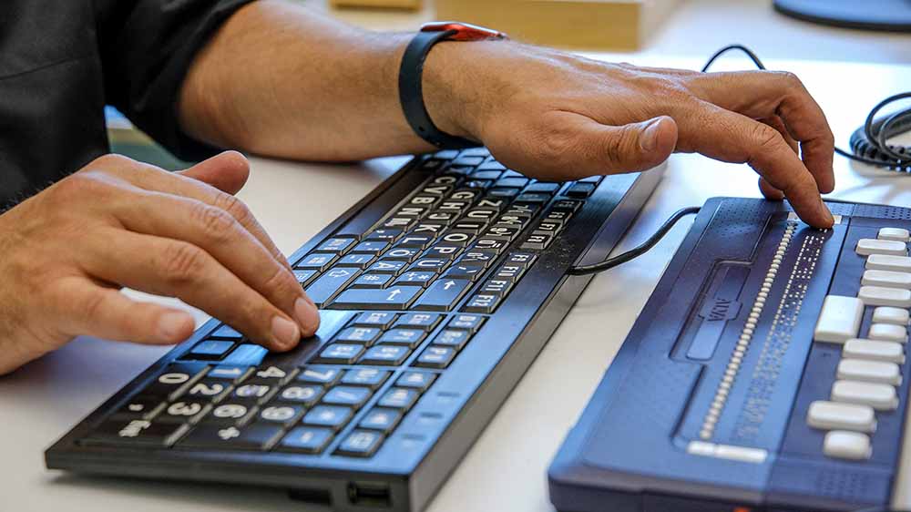 Hand tippt an Tastatur, darüber Vorrichtung mit Blindenschrift