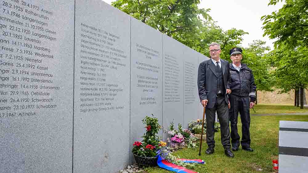 Mann im Anzug mit Stock steht mit einem Mann in Polizeiuniform vor einer Gedenktafel