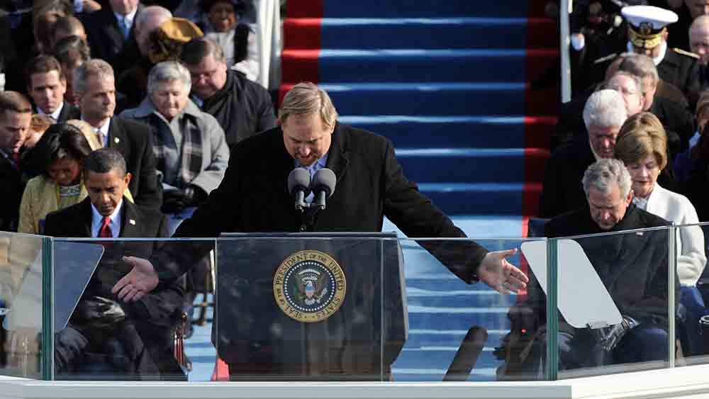 Mann breitet auf Bühne Hände zum Gebet aus, im Hintergrund sitzen George W. Bush und Barack Obama