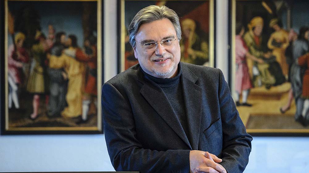 Mann mit schwarzem Sakko und Brille, im Hintergrund hängen Gemälde