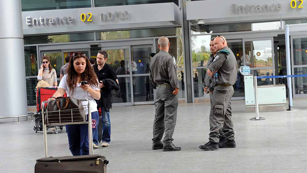 Vor einem Flughafengebäude schiebt eine Frau ihre Koffer, daneben stehen Sicherheitsleute