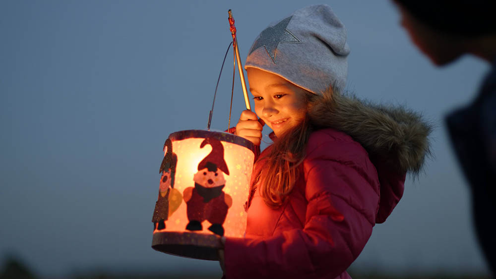 Kind mit grauer Mütze schaut in seine strahlende Laterne und lacht