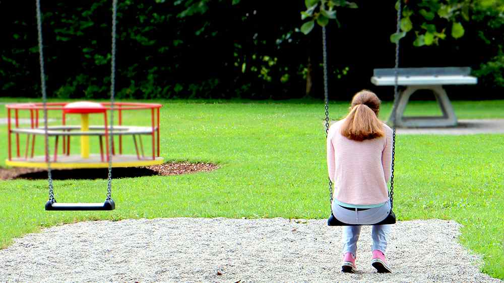 Ein Mensch sitzt leicht vornübergebeugt auf einer Schaukel, auf dem Spielplatz ist sonst niemand.