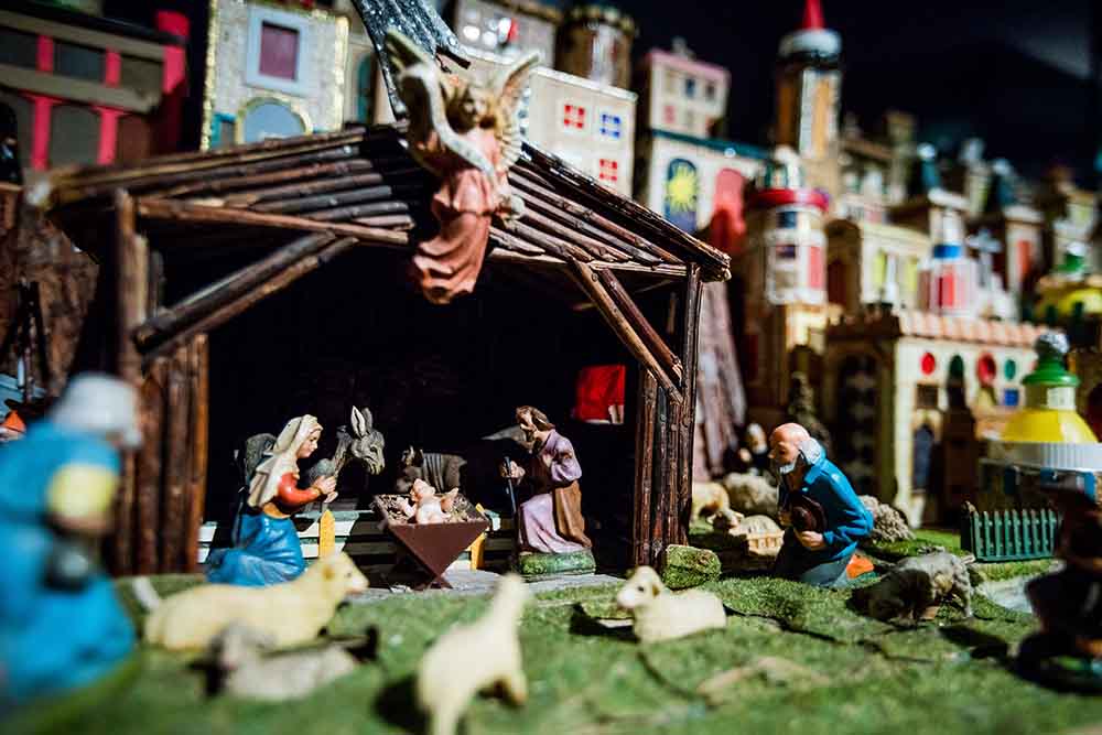 Im Vordergrund ist ein Stall, der aus Ästen gebaut wurde. Ein Engel ist oben befestigt. Maria und Joseph an einer Krippe sind im Stall. Im Hintergrund sind bunte Hochhäuser.