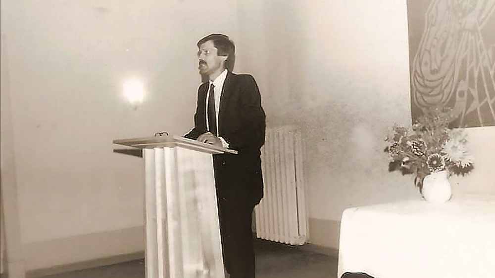 Mann mit Schlips an einem Pult stehend in einem Raum