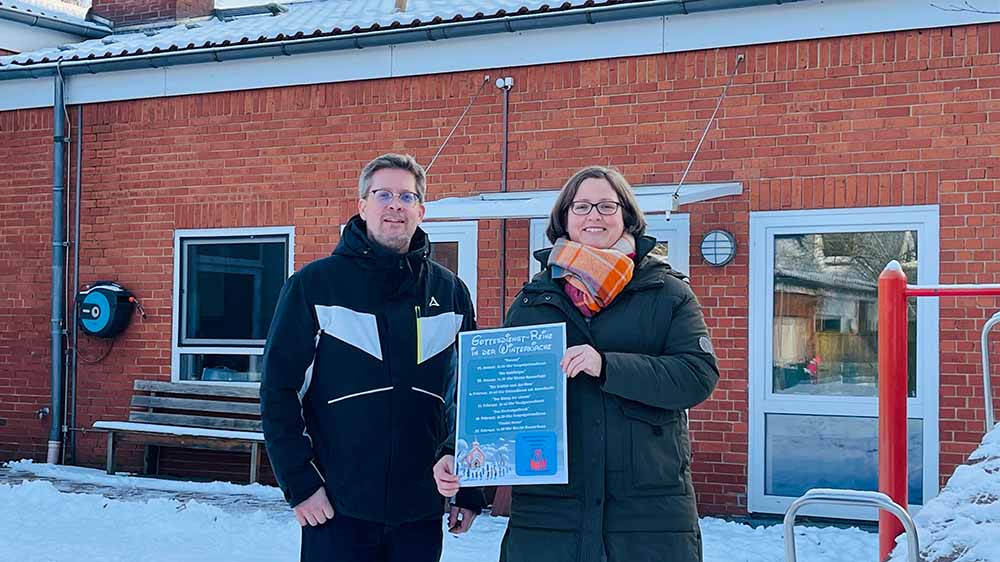 Ein Mann und eine Frau stehen vor einer Hauswand und halten ein kleines Plakat hoch.