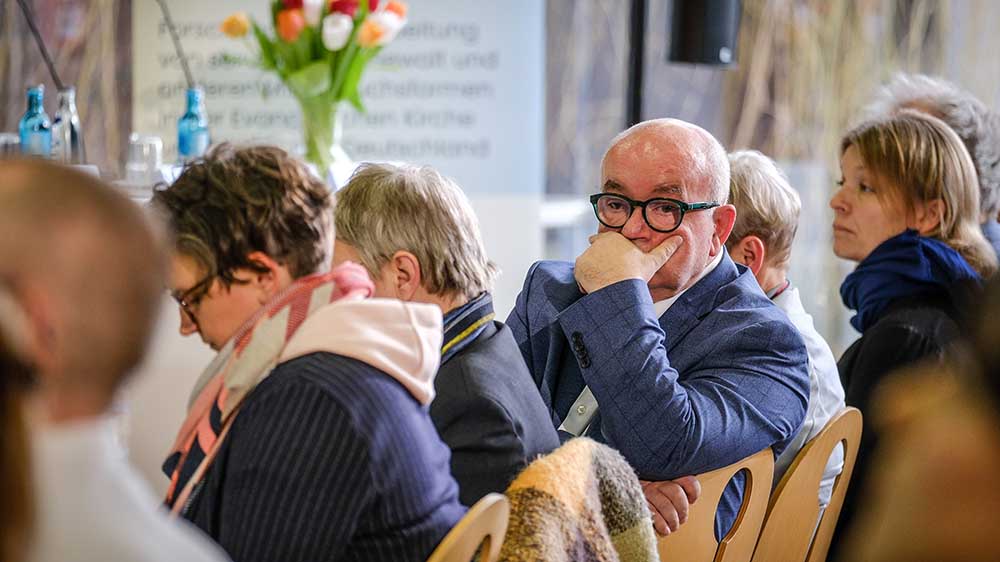 Mann mit Brille sitzt in einer Reihe von Stühlen und halt Hand vor den Mund