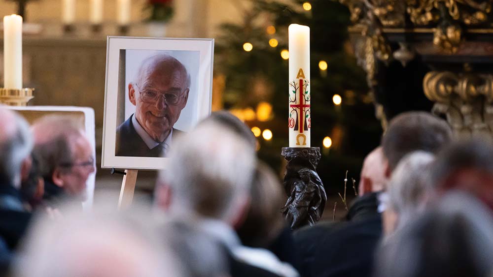 Besucher in einer Kirche, von hinten fotografiert. Vorn steht ein großes Porträtfoto eines älteren Mannes