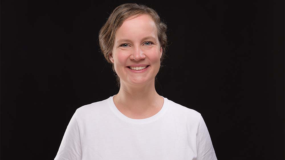 Junge Frau mit dunklen Haaren, die nach hinten geflochten sind. Sie trägt ein weißes T-Shirt.