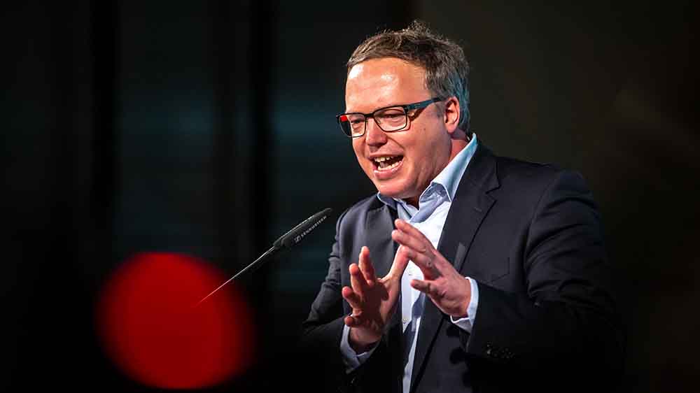 Mann im Sakko und blauen Hemd hält Rede