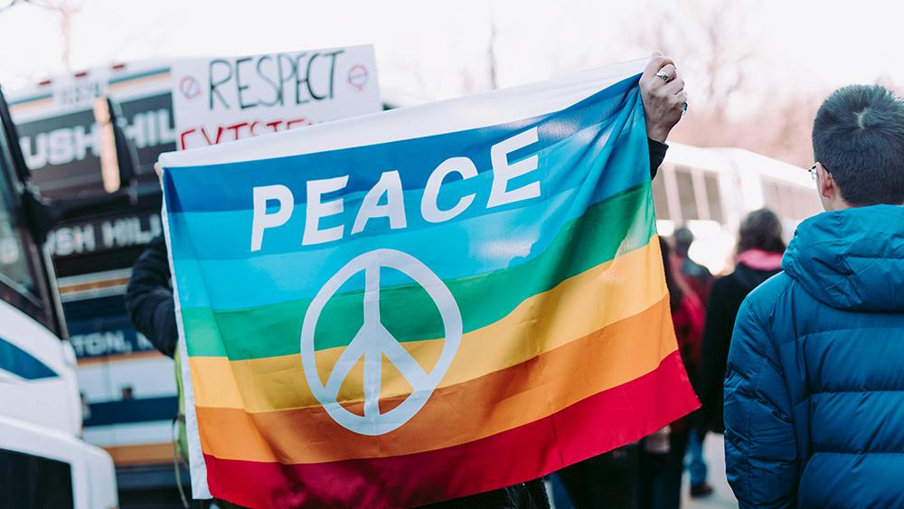 in einer Demonstration hält ein Mensch eine Regenbogenfahne hoch, auf der "Peace" steht.