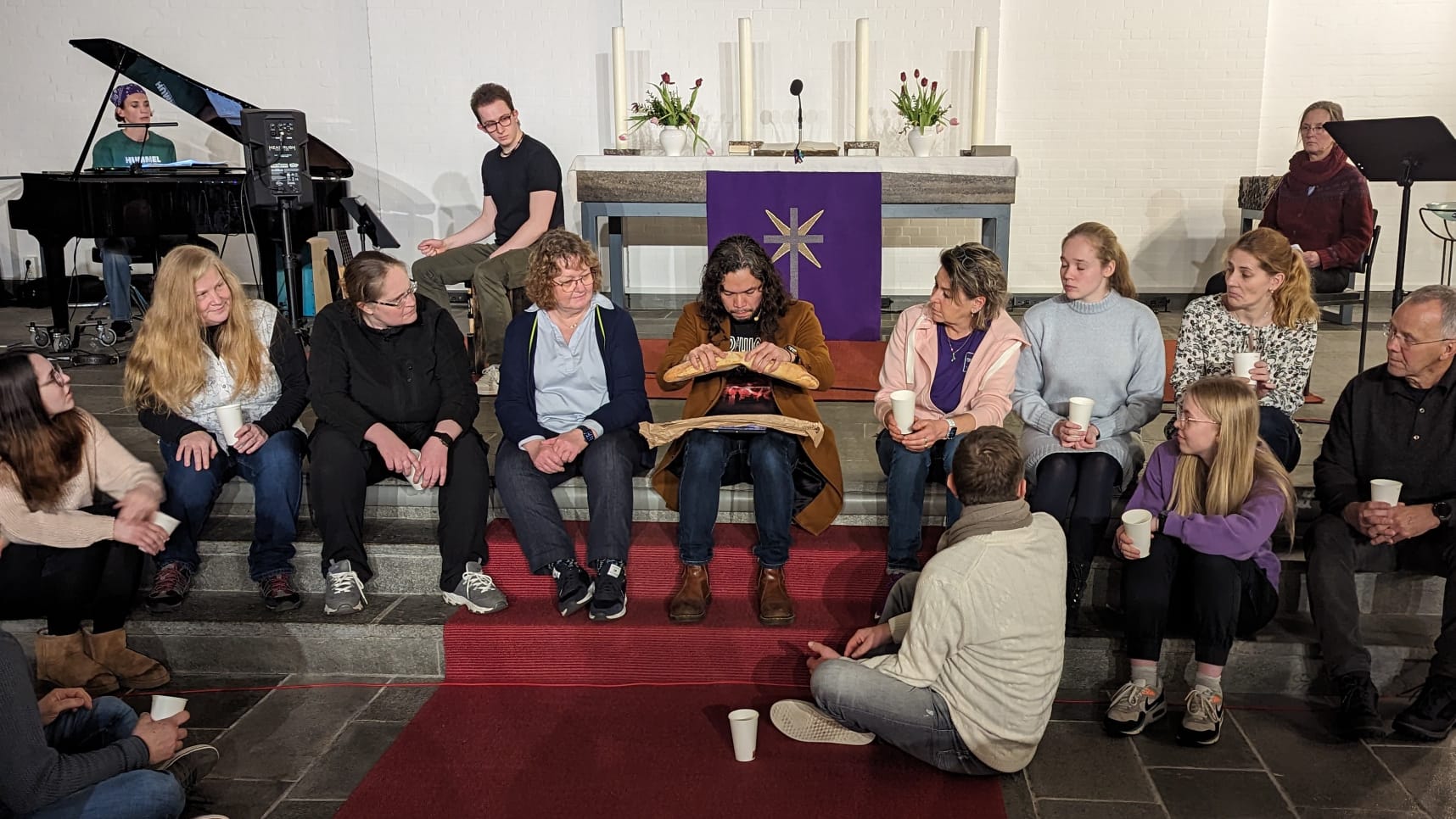 Es ist eine Bild während der Proben zu "Die Passion". Jesus bricht umgeben von Menschen Brot. Im Hintergrund sind Musiker zu sehen.