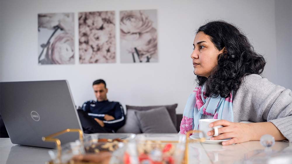 Dunkelhaarige Frau mit lockigem Haar sitzt an einem Tisch mit einem Laptop und einer Tasse. Im Hintergrund sitzt ein Mann auf einem Sofa