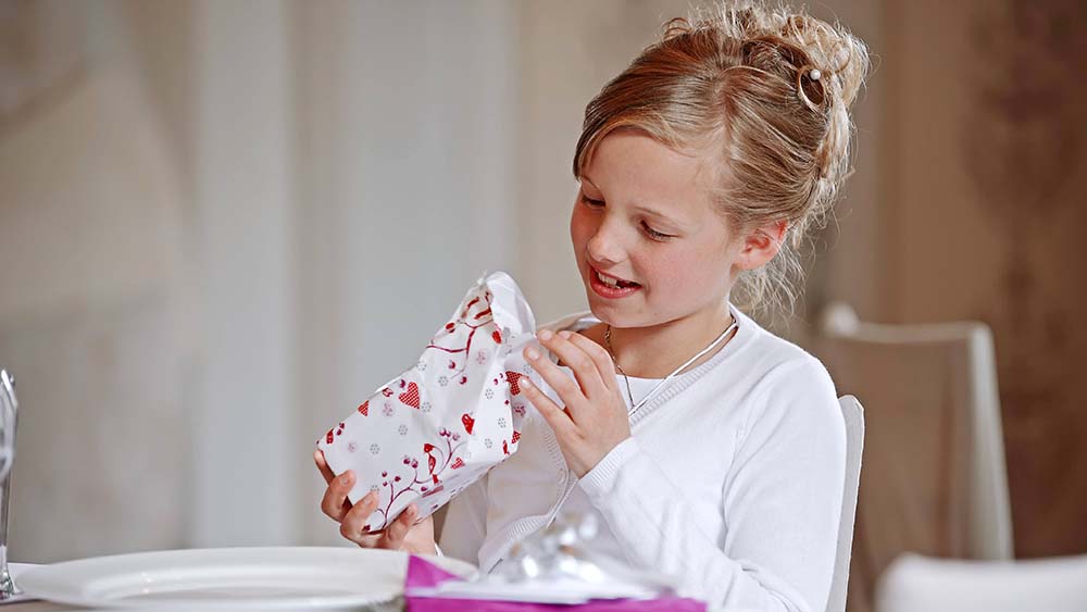 Kleines Mädchen im weißen Shirt sitzt am Tisch und packt Geschenk aus