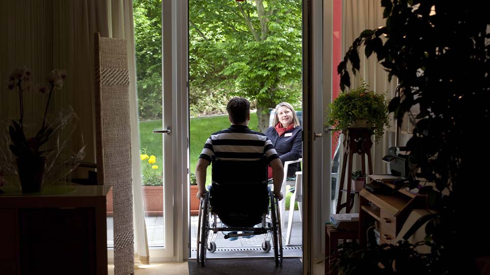 Jüngerer Mann im Rollstuhl fährt auf die Terrasse seiner Wohnung. Dort sitzt eine weibliche Pflegekraft auf einem Stuhl