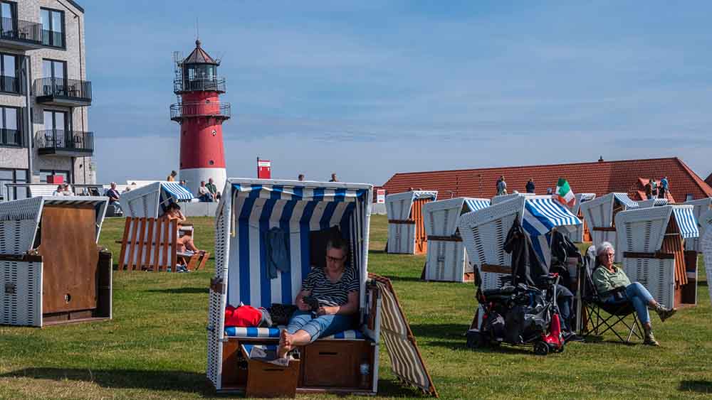 Menschen liegen in Strandkörben an einem Deich, im Hintergrund ein Leuchtturm