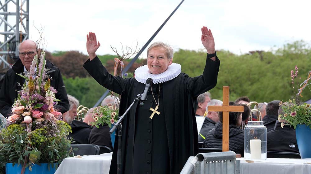 Frau im Talar breitet auf Bühne Arme zum Segen aus