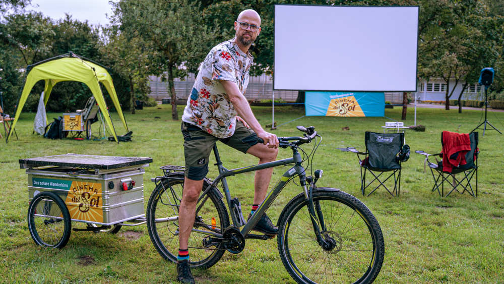 Mann auf Fahrrad mit Anhänger steht auf einer Wiese mit Kinoleinwand