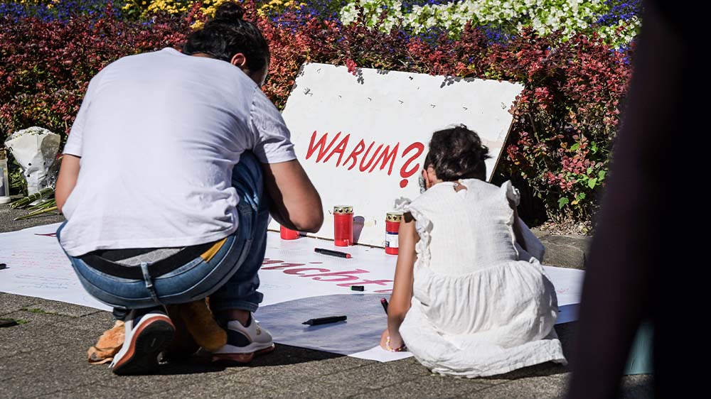 Ein Mann und ein Mädchen sitzen an einem Gedenk-Ort mit Kerzen und einem Plakat mit den Worten "Warum?"