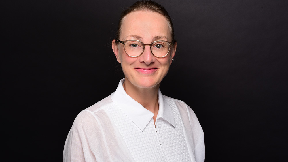 Mittelalte blonde Frau mit zurück gestylten Haaren und einer Brille sowie einer weißen Bluse