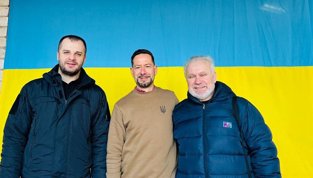 Drei Männer stehen nebeneinander vor einer ukrainischen Flagge und schauen in die Kamera