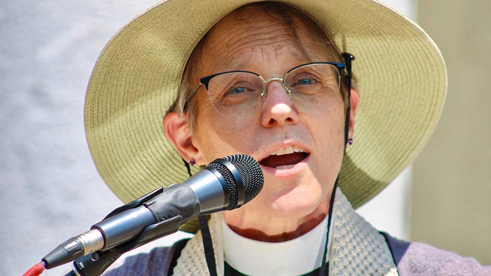 Ältere Frau mit kurzen Haaren und Brille und einem Sonnenhut spricht etwas in ein Mikrofon