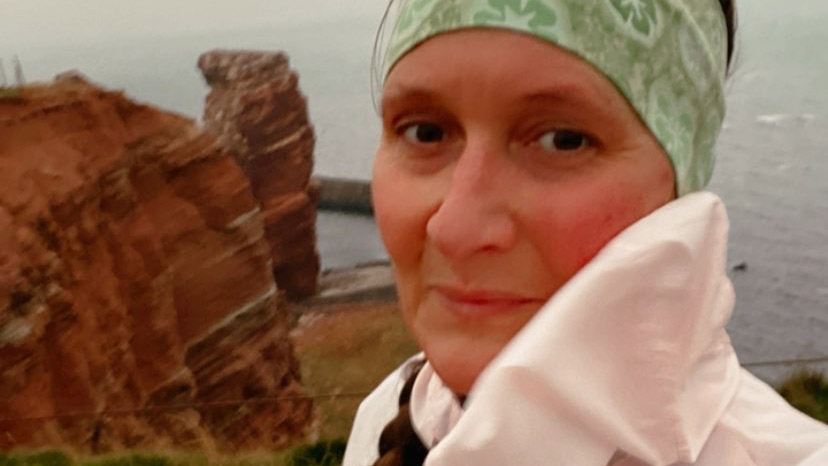 Das Foto zeigt ene Frau in einer zartrose Jacke. Im Hintergrund ist die "Lange Anna" zu sehen. Der Felsvorsprung ist das Wahrzeichen der Insel Helgoland.