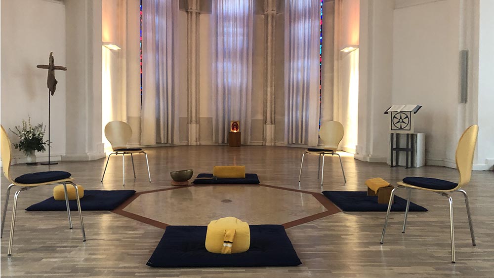 Wenige Stühle, baue Sitzmatten und Meditationskissen bilden einen kleinen Kreis. Der Raum ist hell beleuchtet. An der Wand links hängt ein Kreuz.
