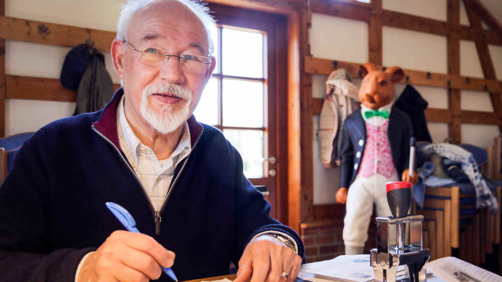 Ältere Mann sitzt mit Stift in der Hand am Schreibtisch