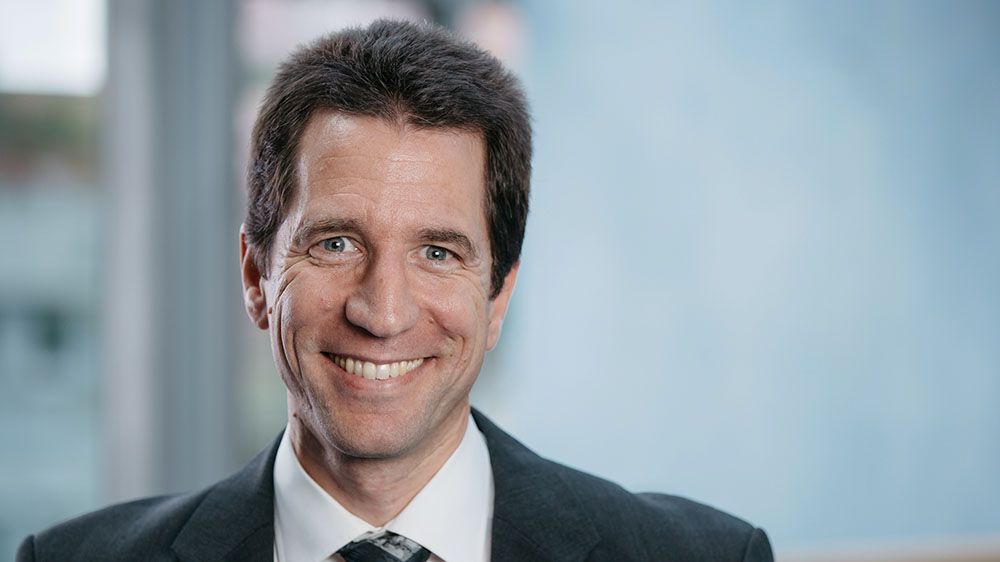 Ein Mann mittleren Altern im Anzug lächelt in die Kamera. Der Hintergrund des Bildes ist unscharf.