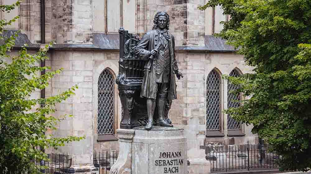 Eine Bronzestatue von Johann Sebastian Bach in Lebensgröße im Mittelpunkt des Fotos. Im Hintergrund sind Kirchenmauern zu sehen und Bäume.