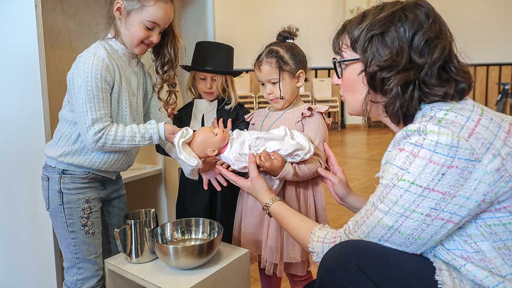 Drei kleine Kinder halten eine Puppe in der Hand und schütten Wasser drüber, eine Frau kniet daneben und passt auf