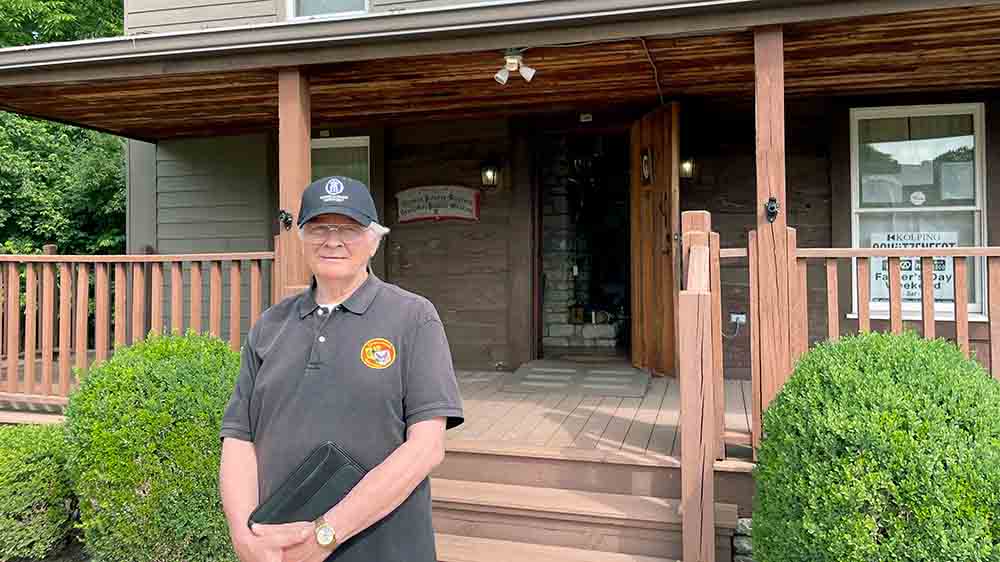 Ein älterer Mann mit Baseballcap steht vor einem Haus und schaut in die Kamera.