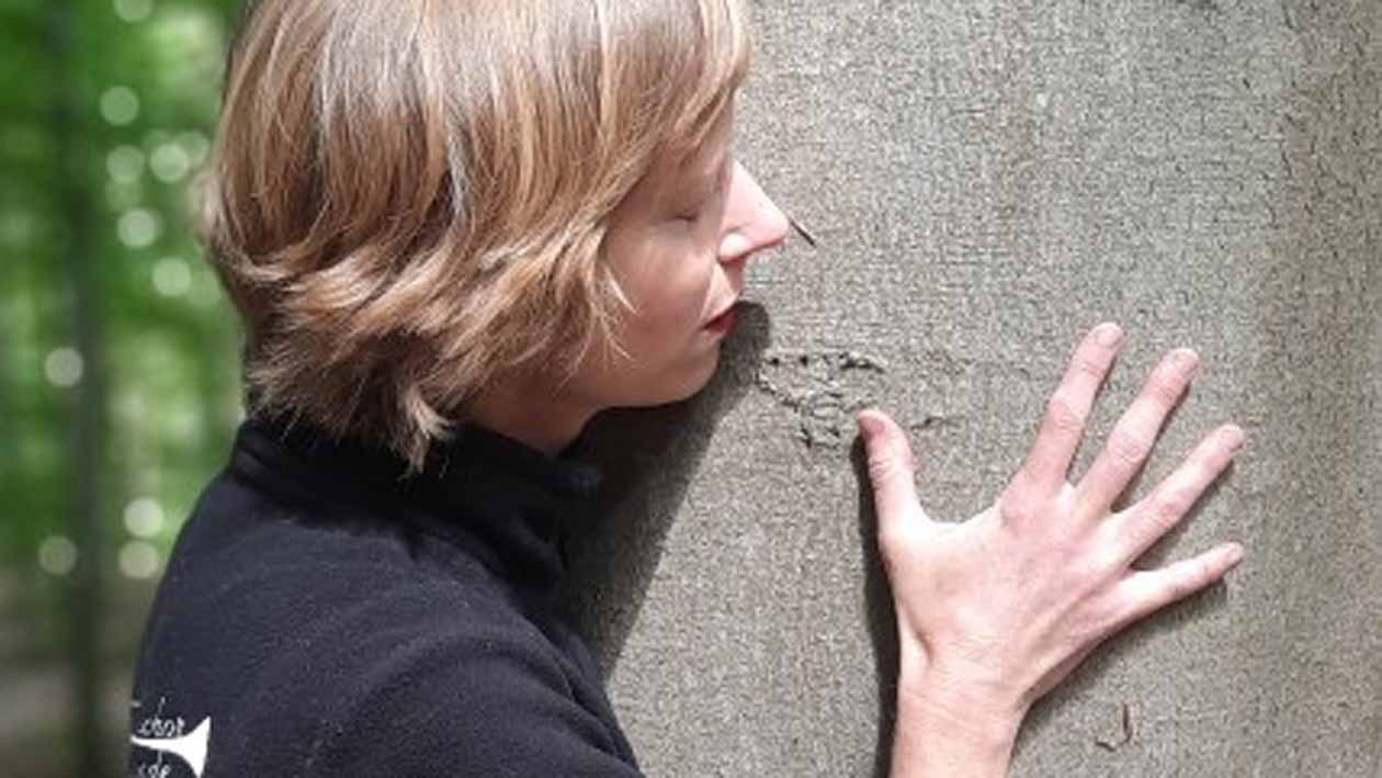 Eine Frau mit dunkelblondem kurzen Haar hält ihr Gesicht und ihre rechte Handfläche an einen grauen glatten Baumstamm