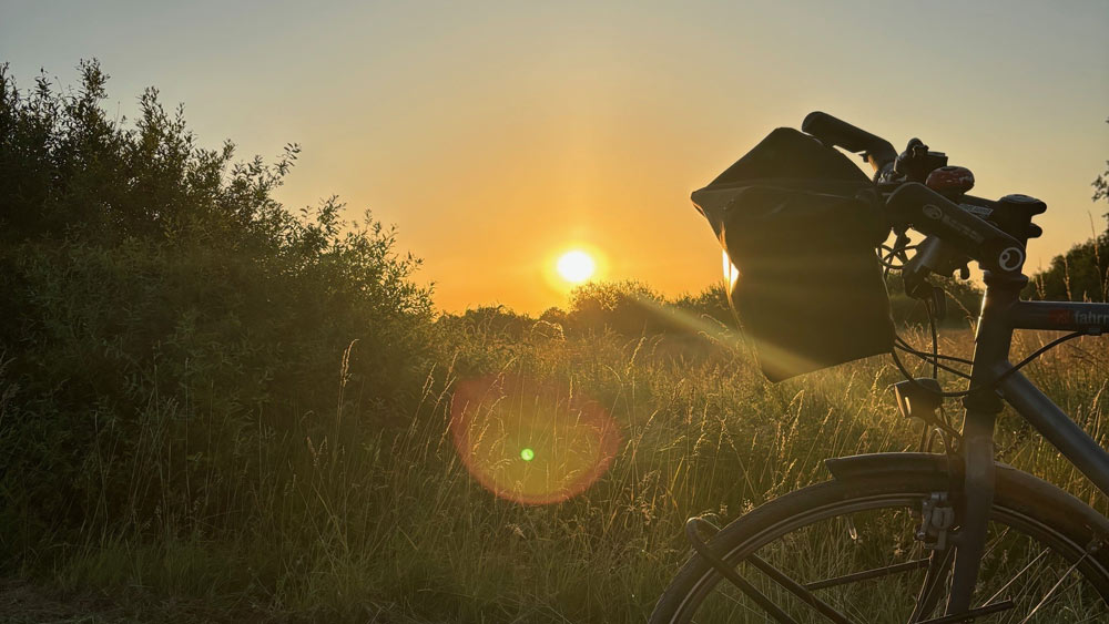 In der Mitte des Bildes eine orangefarbene Sonne kurz nach dem Aufgang. Rechts ein Fahrradlenker, links ein Busch.