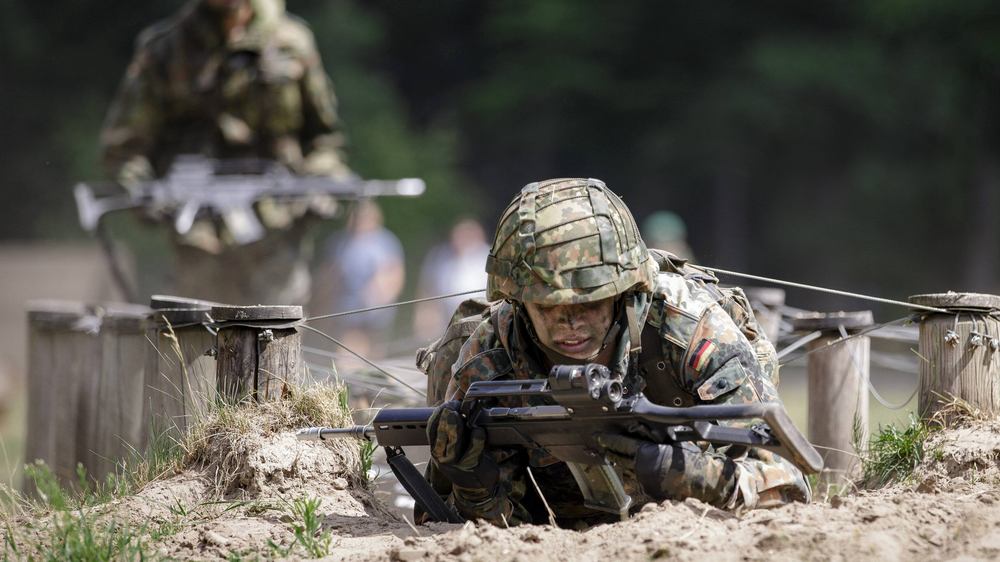 Soldat im Kampfmontur robbt mit Gewehr über ein sandiges Feld. Weiterer Soldat im Hintergund schaut zu