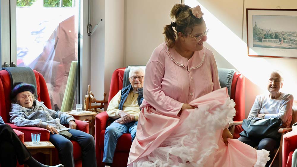 Eine Frau mit einem rosafarbenen Tüllkleid lächelt und tanzt. Hinter ihr sitzen ältere Menschen, die lächeln