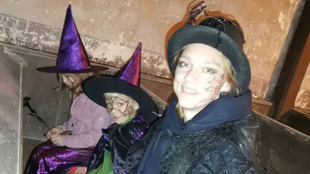 Ein Kind, das als Zauberer verkleidet ist, und zwei als Hexen verkleidete Kinder, sind vor einem alten Gemäuer zu sehen