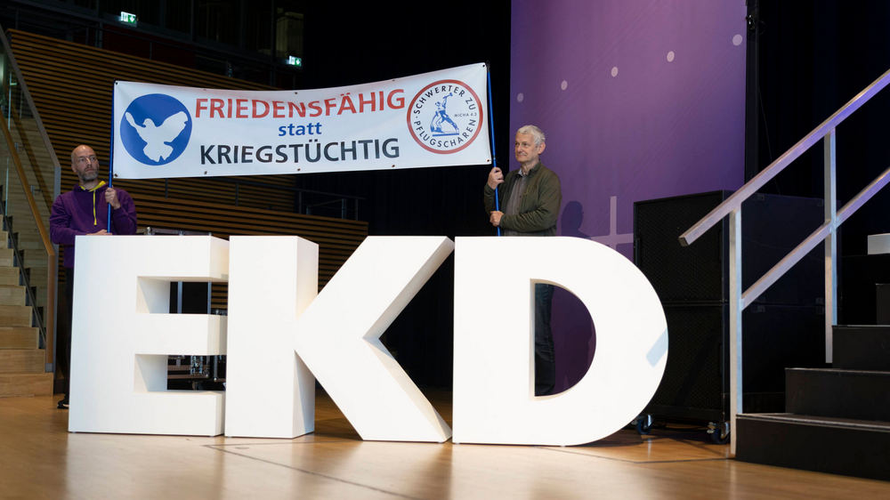 In einem Tagungssaal stehen zwei Männer hinter den Buchstabend "KED". Sie halten ein Schild hoch, auf dem steht: Friedensfähig statt kriegstüchtig
