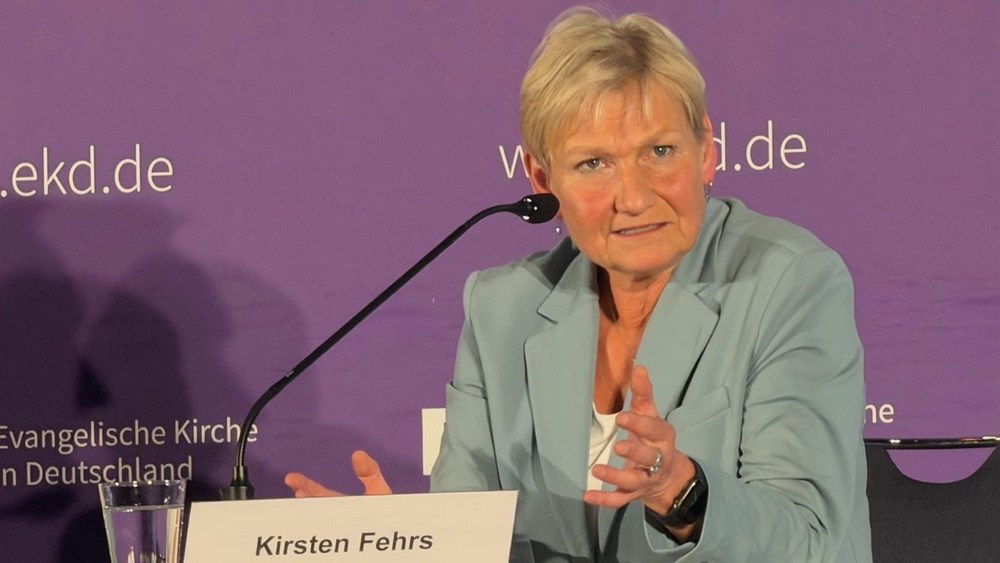 Frau im grünen Blazer und blonden Haaren sitzt auf Podium und macht Geste mit linker Hand