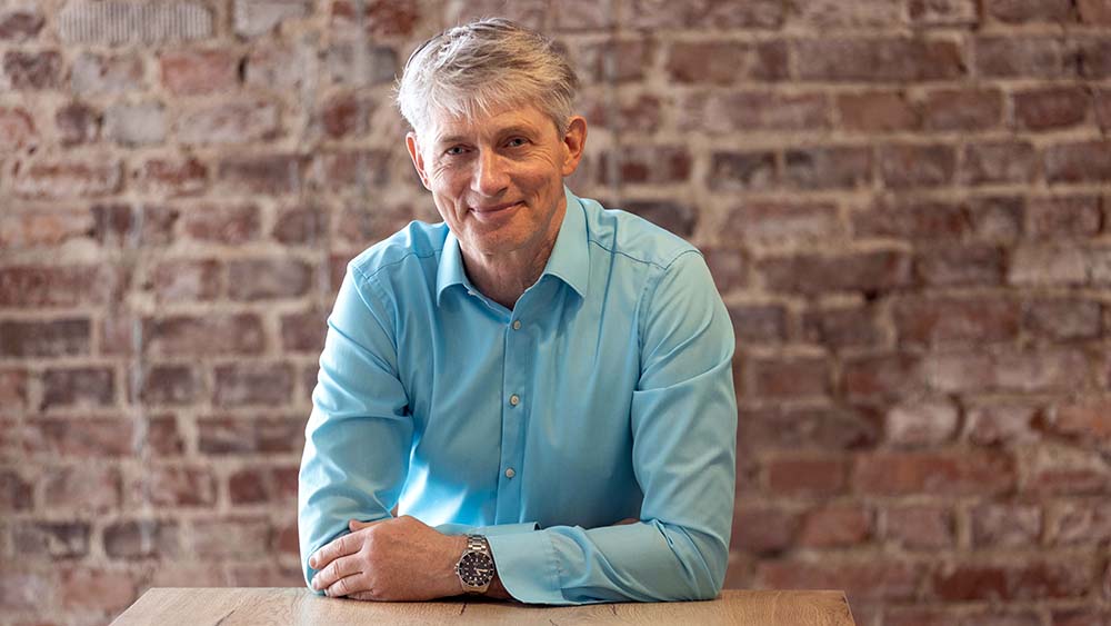 Mann im grünen Hemd und mit grauen Haaren lehnt auf einem Holztisch, Backsteinwand im Hintergrund