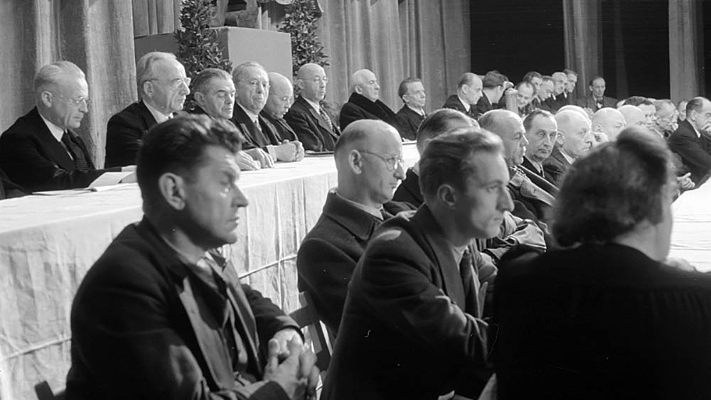 Ein Schwarz-Weiß-Foto von einer Sitzung. Zu sehen sind Männer an Tischen mit weißen Tischdecken.