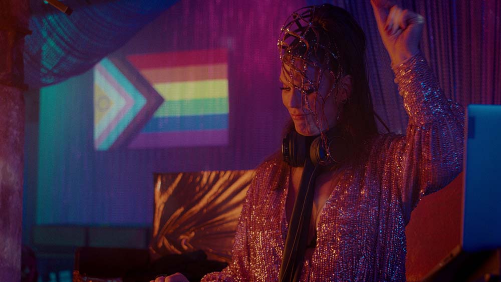 Eine dunkle Szene in einem CLub. Eine Frau steht mit einem erhobenen Arm und mit Kopfschmuck im Fokus und sieht nach unten. Im Hintergrund eine Regenbogenflagge.