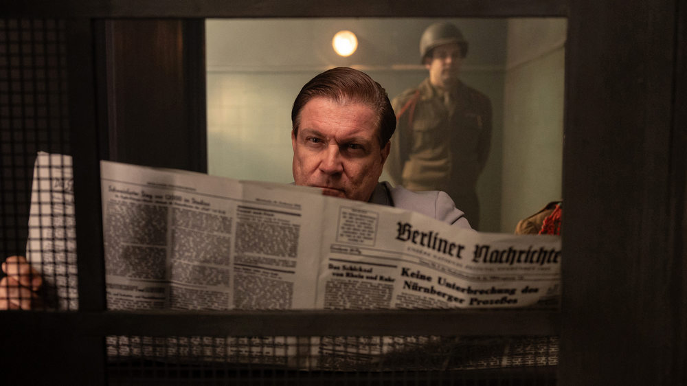 Mann liest in einer Zeitung, im Hintergrund steht ein Soldat in Uniform