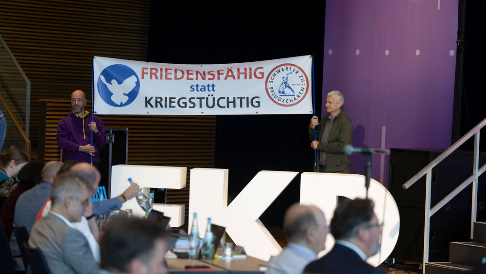 In einem Tagungssaal stehen zwei Männer hinter den Buchstabend "KED". Sie halten ein Schild hoch, auf dem steht: Friedensfähig statt kriegstüchtig