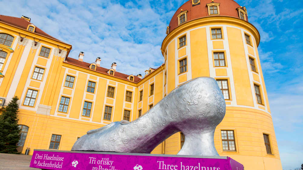 Gelbes Barockschloss mit einem riesigen silbernen Schuh auf einem pinken Podest