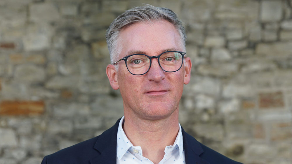 Ein mann mit grauem Haar und Brille steht vor einer Steinwand und schaut in die Kamera.