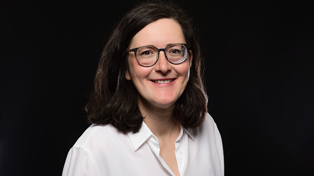 Eine Frau mit dunkeln Haaren, weißer Bluse und einer dunklen Brille lächelt in die Kamera.